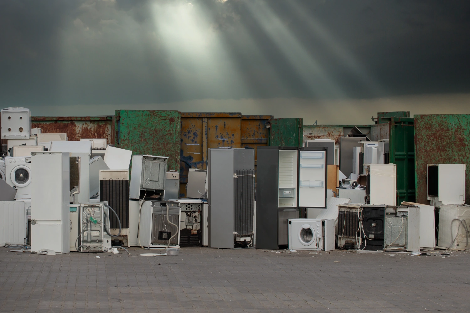 A variety of discarded household appliances, including refrigerators and washing machines, gathered for recycling at an outdoor facility under a dramatic sky.