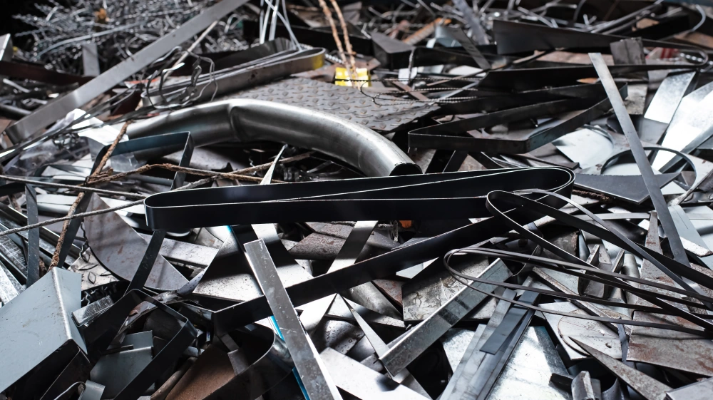 A variety of steel scrap, including flat strips, curved metal piping, and textured floor plates, piled at an industrial recycling yard.