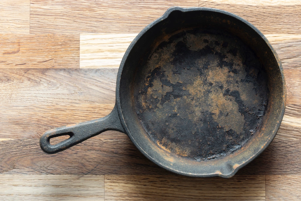 Overhead view of a heavily rusted and charred cast iron skillet resting on a light-colored wooden surface.