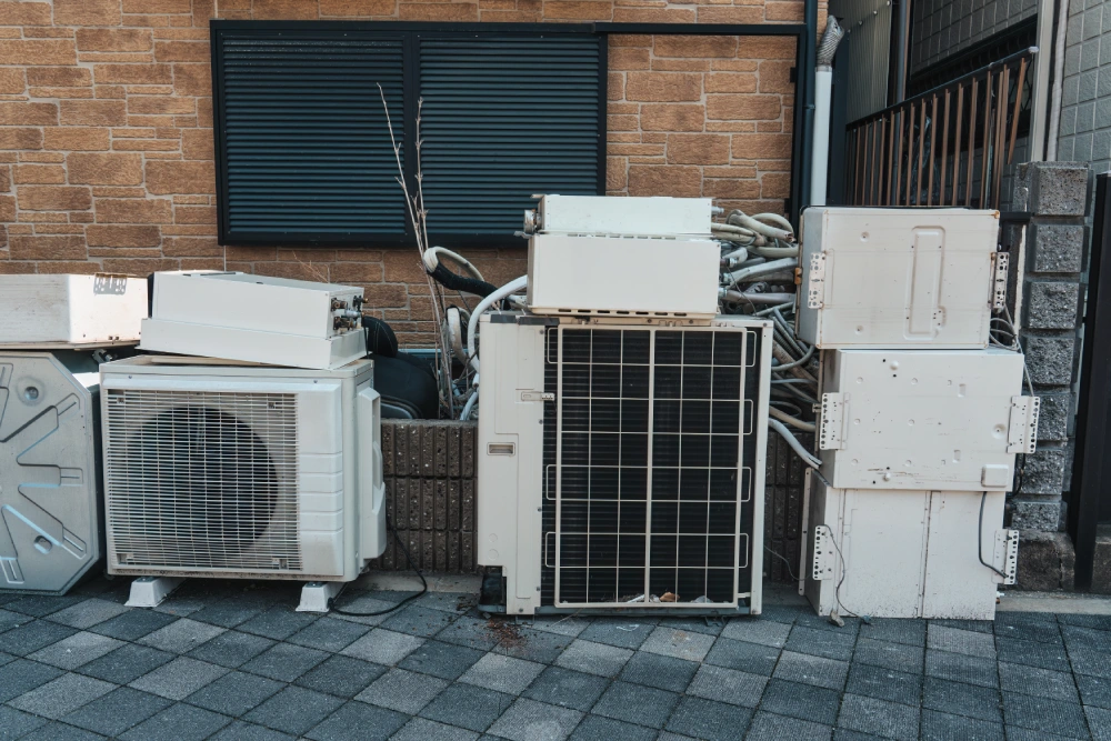 A collection of old air conditioning condenser units and metal HVAC components stacked outside a building for recycling.