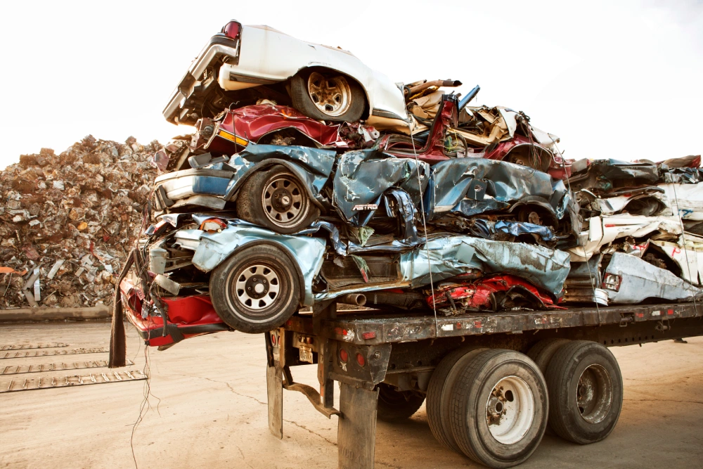 Several crushed and flattened cars are stacked high on the back of a large flatbed trailer in a scrap yard, ready for transport and final processing.