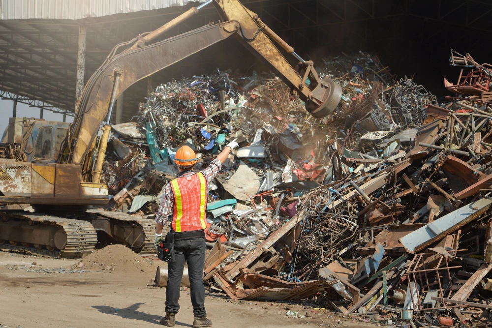 A recycling yard worker in a safety vest signals to an excavator operator moving a large pile of scrap metal for processing.