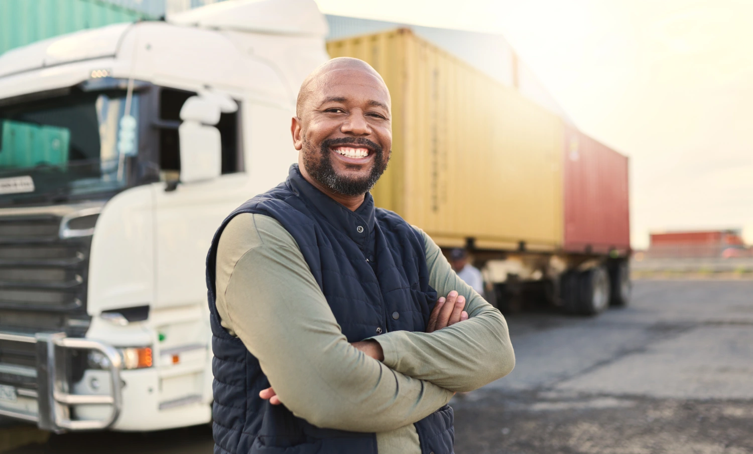 A smiling transport truck driver stands with arms crossed in front of a white semi-truck and yellow shipping containers.