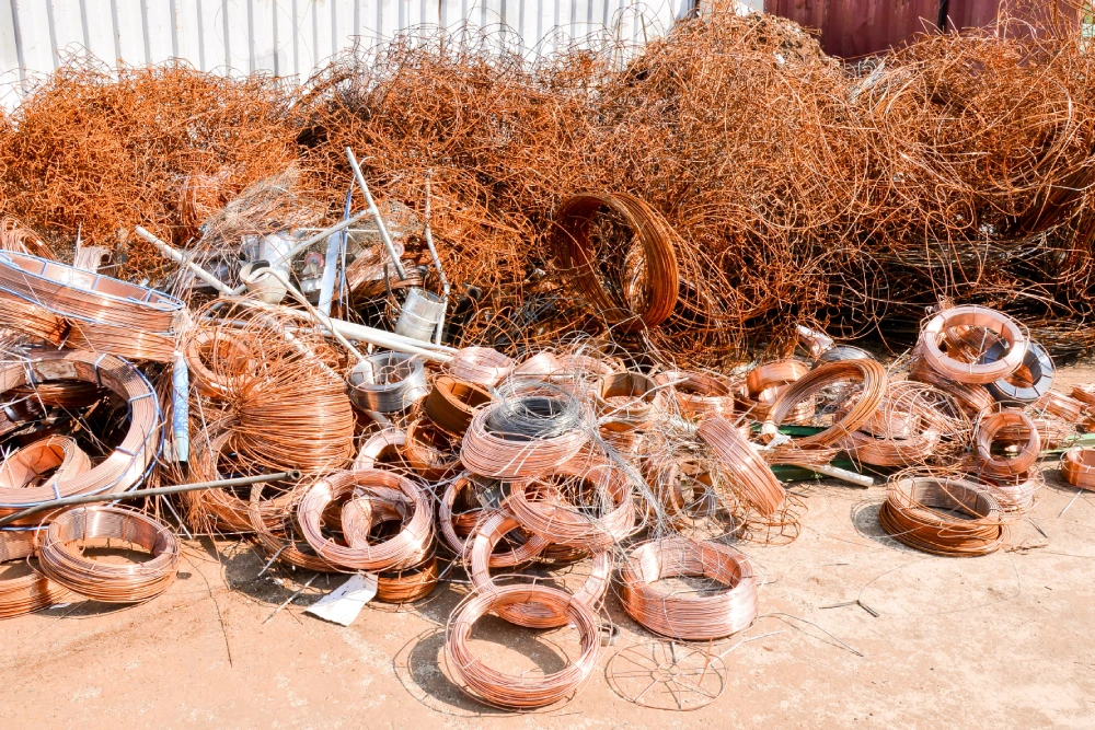 A variety of scrap copper materials, including shiny coils of new copper wire and large tangles of weathered, dark copper wiring, sorted on a concrete floor at a recycling center.